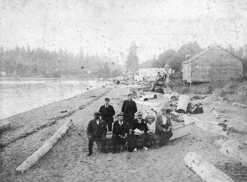 1890?-View of English Bay beach looking west-Photograph shows Joe Fortes seated on the far left and Roderick F. MacKenzie standing behind boat on the right