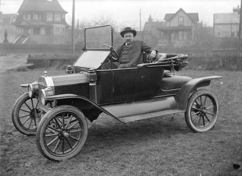 1919-Man seated in car parked in clearing near houses