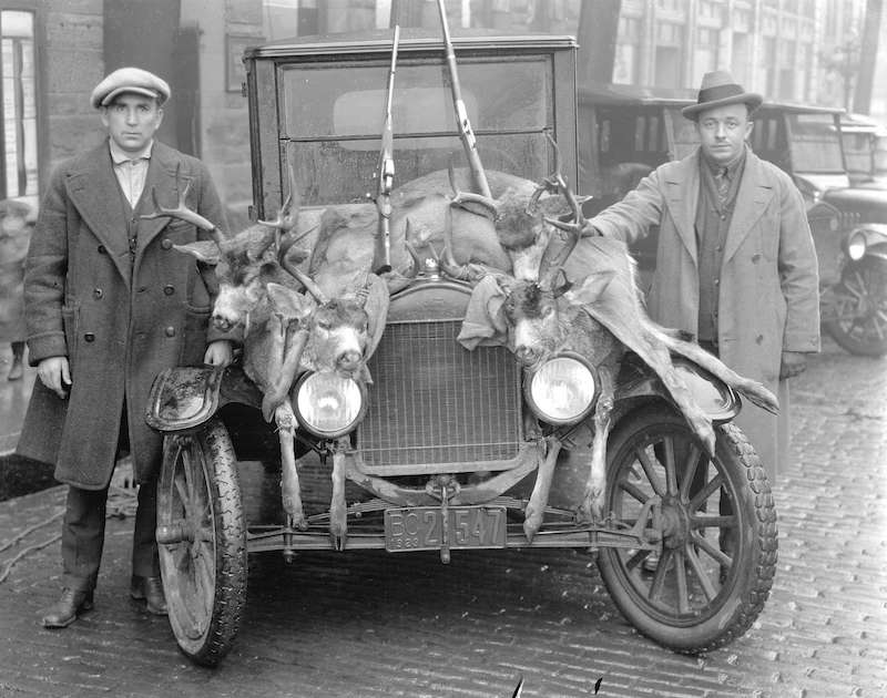 1923-Two men posing with dead deer draped over the hood of a car