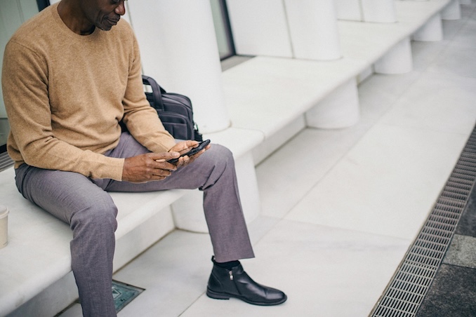 man using a cell phone while sitting on a ledge outside.