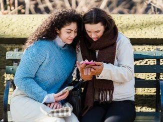 two women on a bench in a park using their cellphone for featured image