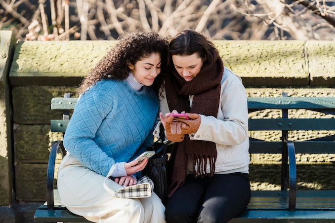 two women on a bench in a park using their cellhone